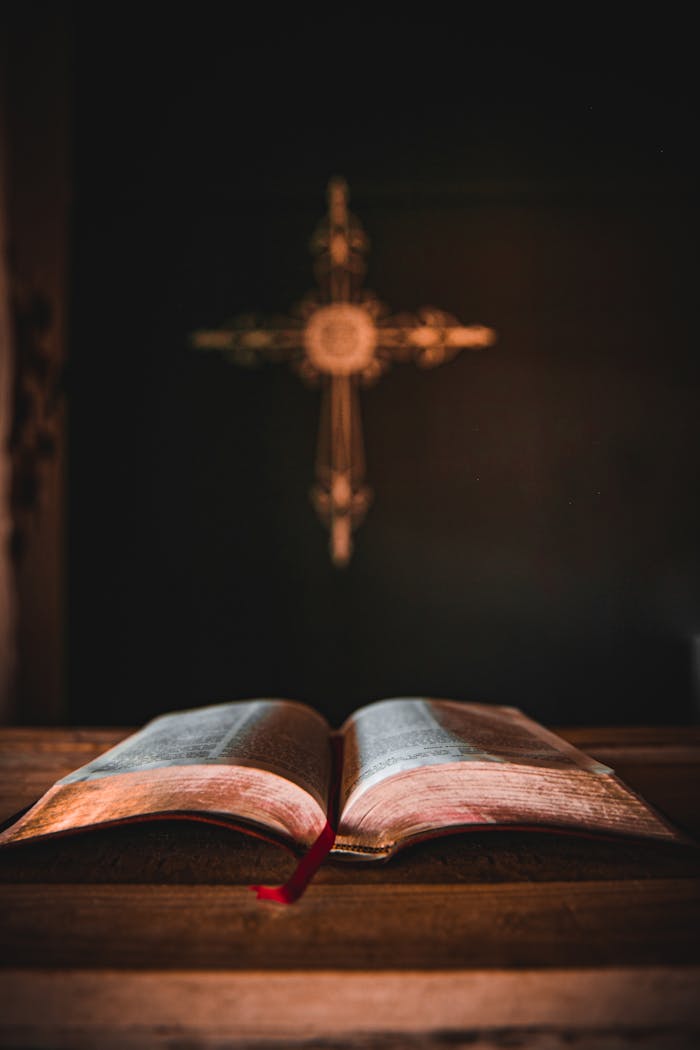 An open Bible on a wooden table with a cross in the background, suggesting a spiritual theme.